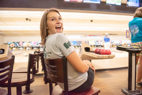 A woman sitting in a chair laughs while people in background bowl at bowling alley